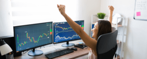 women in front of 2 computer screens showing stock charts graphs